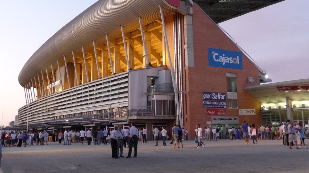 Fachada exterior del estadio Nuevo Colombino con publicidad de Cruzcampo y Cajasol al atardecer, con aficionados en la explanada.