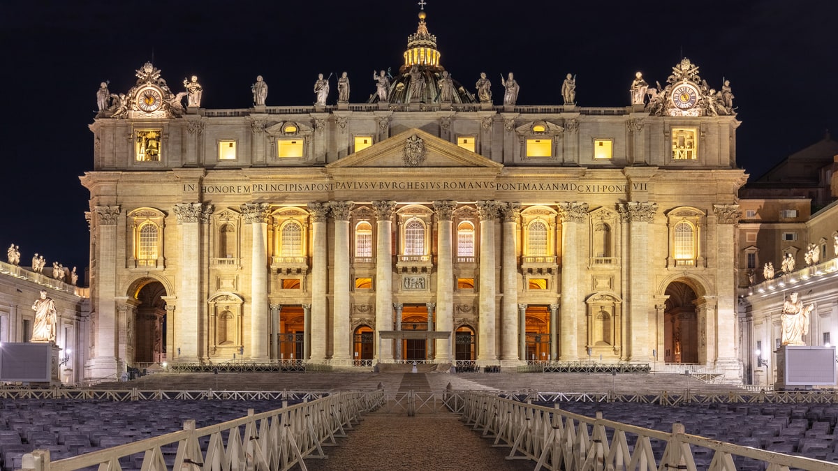 Fachada de la Basílica de San Pedro iluminada de noche, con la plaza vacía y sillas al frente.