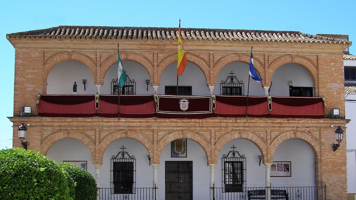 Fachada del Ayuntamiento de Bollullos Par del Condado con arcos de ladrillo, banderas y bancos en la plaza