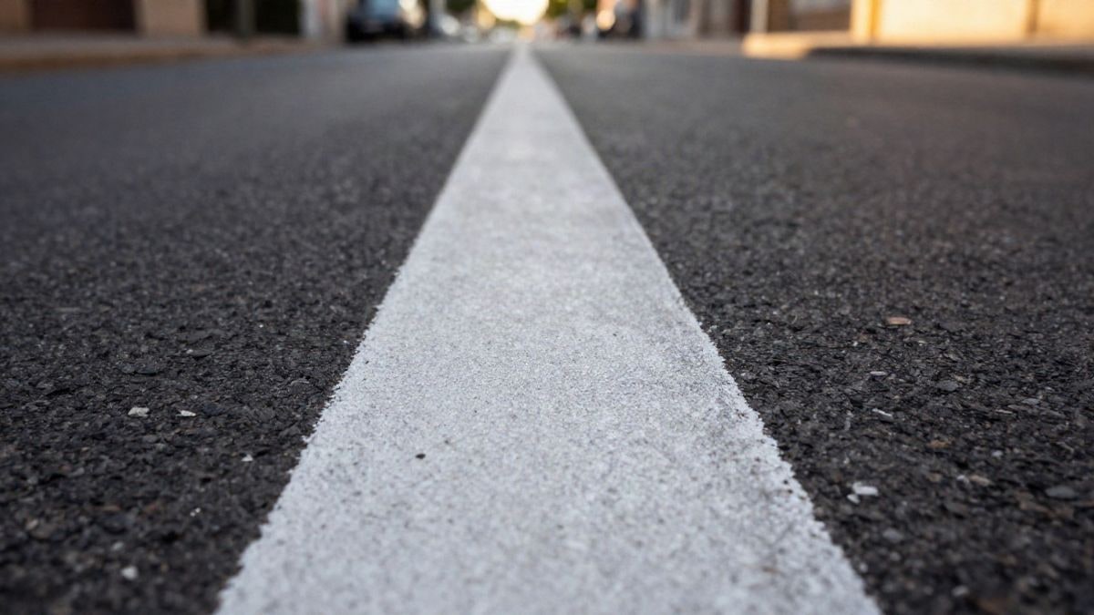 Imagen ilustrativa: línea blanca de señalización recién pintada sobre el asfalto al atardecer, con una calle desenfocada al fondo