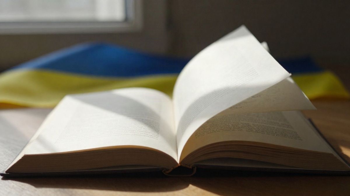 Imagen ilustrativa: un libro abierto sobre una mesa de madera con luz natural y una cinta con los colores de Ucrania al fondo
