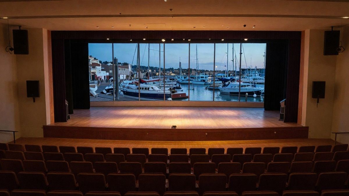 Imagen ilustrativa: un auditorio vacío con un escenario iluminado y ventanales que dejan ver un puerto al atardecer.
