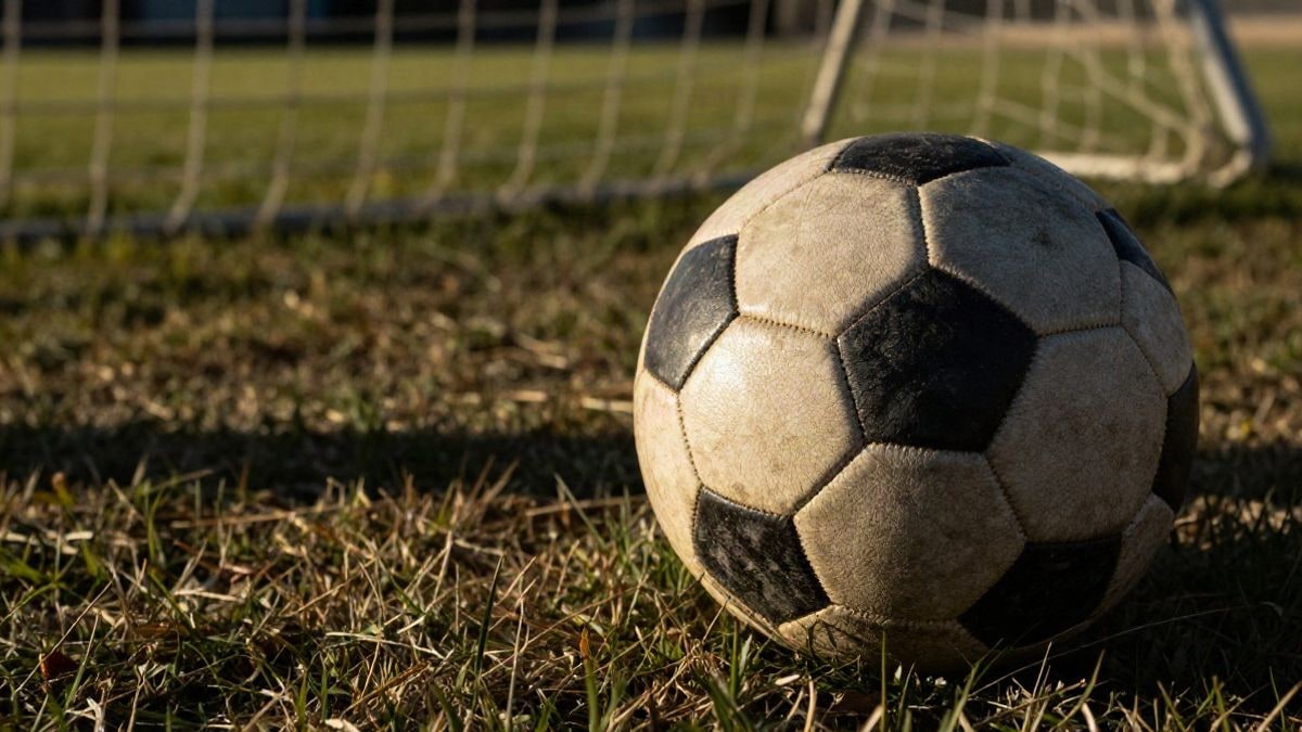 Imagen ilustrativa: detalle de un balón de fútbol desgastado sobre hierba seca con una portería desenfocada al fondo