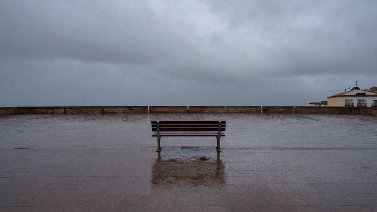 Imagen ilustrativa: un banco de madera mojado en una plaza vacía de una ciudad española durante una lluvia al atardecer