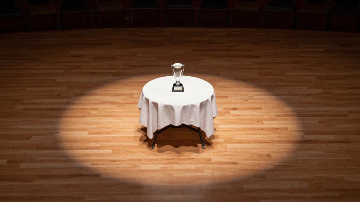 Imagen ilustrativa: un trofeo de cristal sobre una mesa cubierta de blanco en un escenario vacío, iluminado por un foco suave