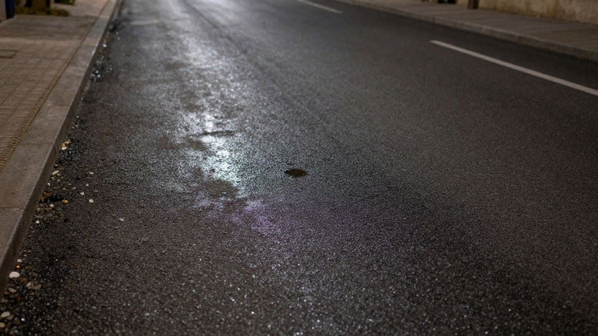 Imagen ilustrativa: carretera asfaltada vista desde arriba de noche, con grava y un charco reflejando la luz de una farola
