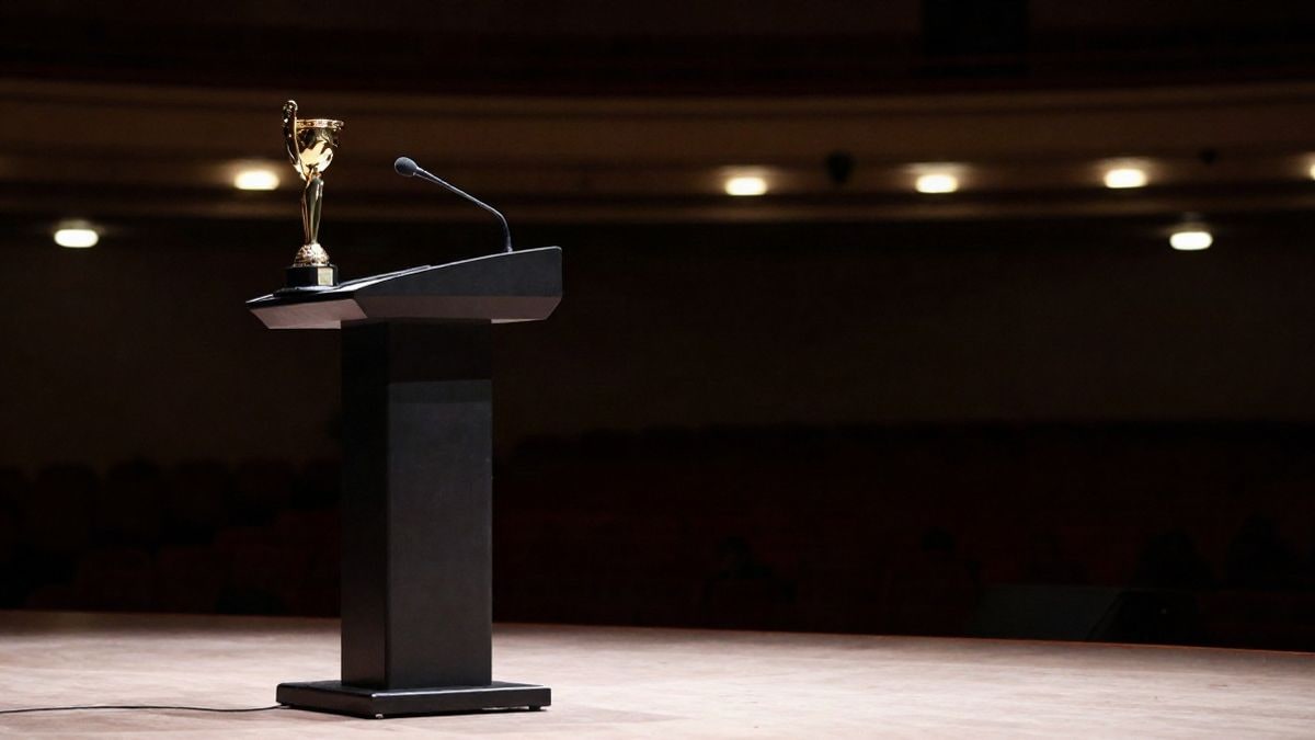 Imagen ilustrativa: un podio con un trofeo dorado y un micrófono en un auditorio con luces de fondo