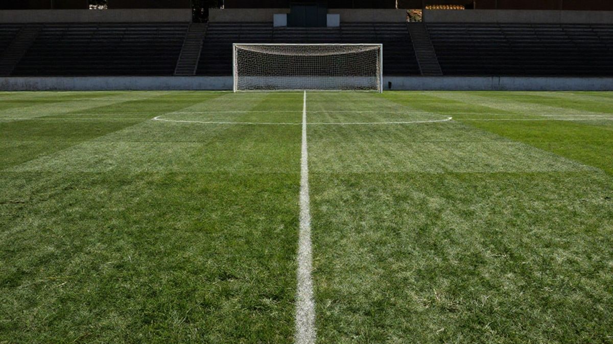 Imagen ilustrativa: campo de fútbol modesto con líneas que convergen hacia la portería y gradas vacías al fondo