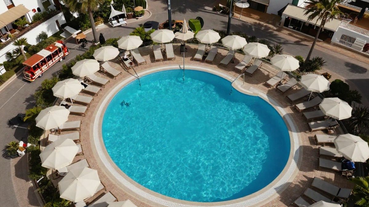 Imagen ilustrativa: vista cenital de una piscina de hotel con tumbonas y un tren turístico en un entorno costero