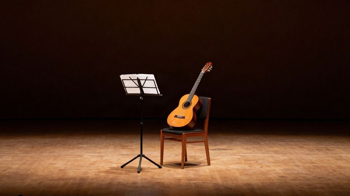 Imagen ilustrativa: escenario de un auditorio con una silla vacía y una guitarra española junto a un atril