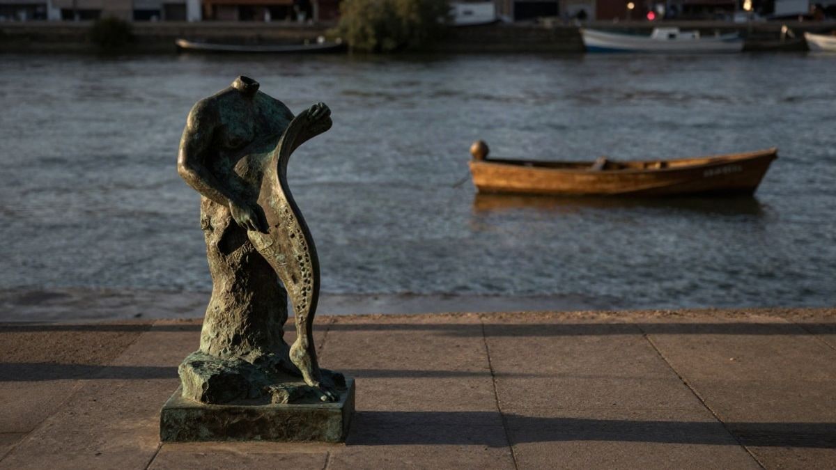 Imagen ilustrativa: pedestal de piedra junto a un paseo fluvial al atardecer, con barcas de madera desenfocadas al fondo