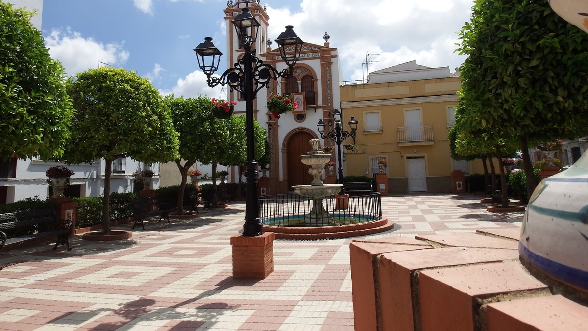 Plaza de Bonares con fuente, farolas negras, iglesia de fachada blanca y árboles recortados