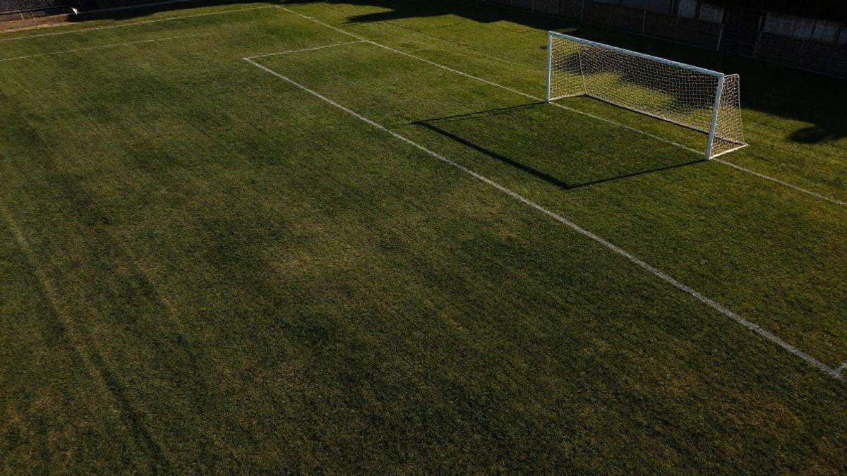Imagen ilustrativa: vista cenital de un campo de fútbol local con líneas blancas y portería, iluminado por el sol de la tarde