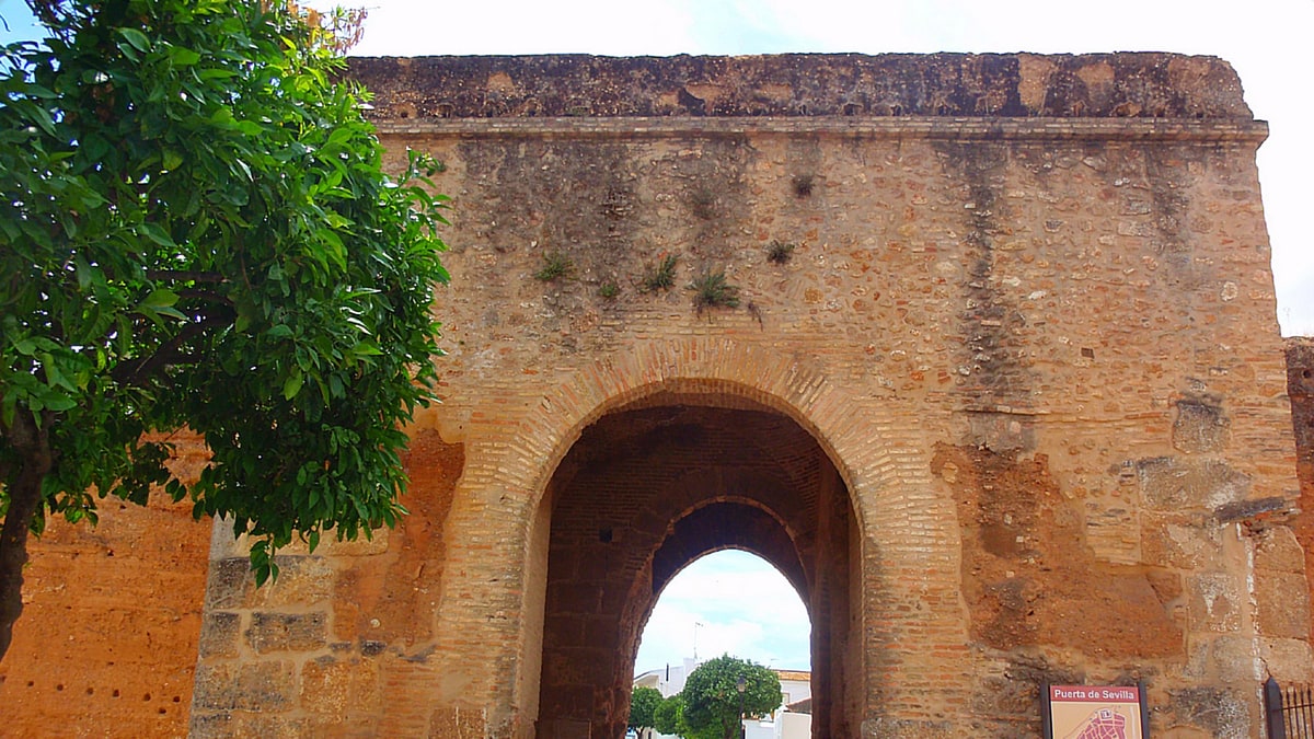 Puerta de Sevilla de Niebla, arco de ladrillo y piedra con calle y coches al fondo.