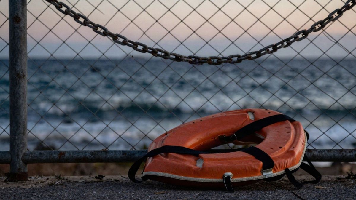 Imagen ilustrativa: detalle de una valla metálica desgastada junto al mar al atardecer con un chaleco salvavidas en el suelo