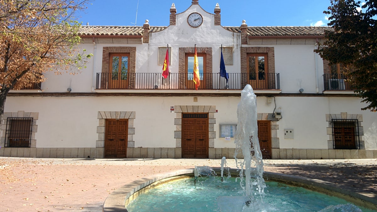 Fachada del Ayuntamiento de Urda con banderas, reloj y una fuente en primer plano