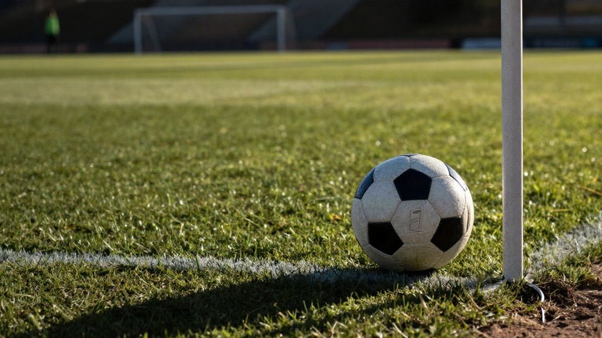 Imagen ilustrativa: un balón de fútbol sobre un césped natural desigual, con una esquina del campo y gradas desenfocadas al fondo.
