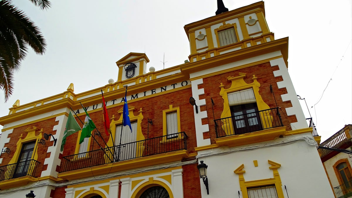 Fachada blanca y ocre del Ayuntamiento de Valverde del Camino con banderas y balcones, a la luz del día