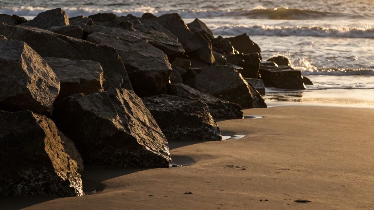 Imagen ilustrativa: un talud de grandes rocas junto a la orilla al atardecer, con sombras largas sobre la arena