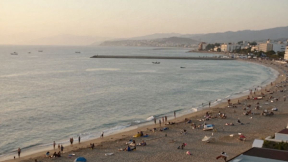 Imagen ilustrativa: vista aérea de la costa mediterránea al amanecer, con una playa concurrida y barcas en la lejanía, sin rostros reconocibles.