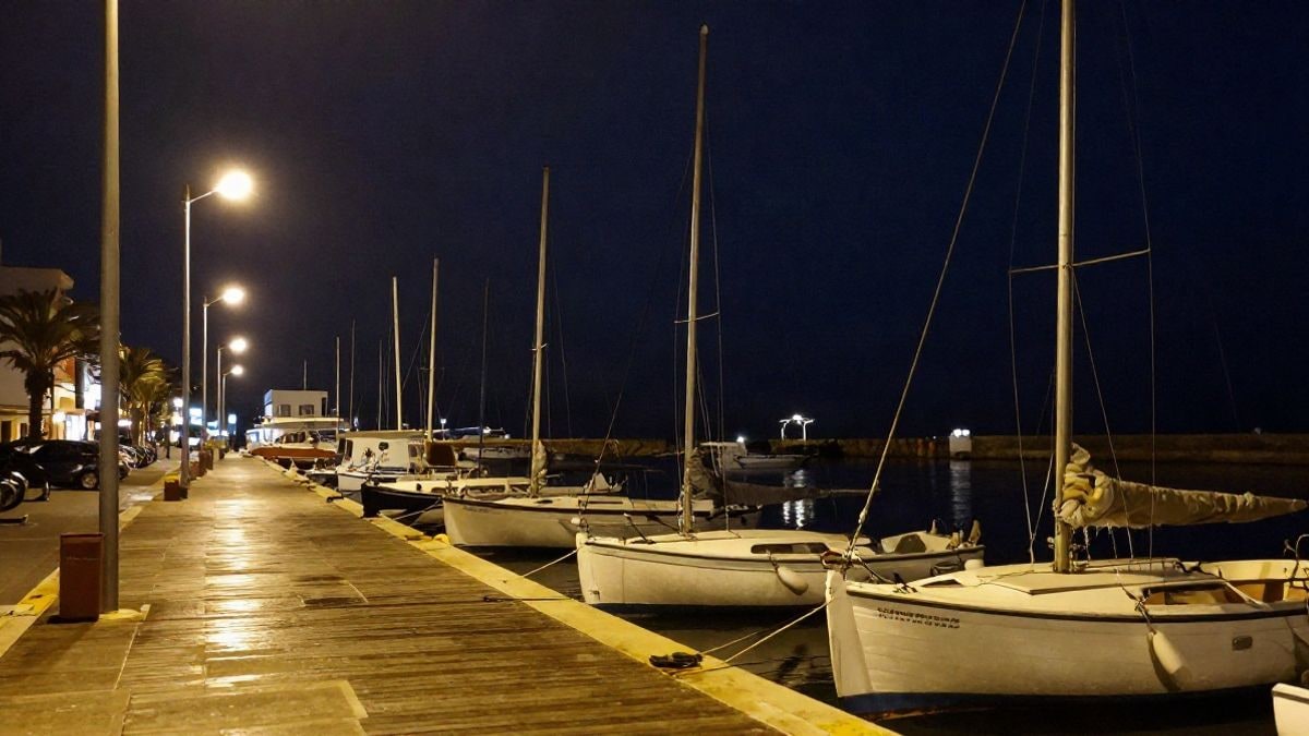 Imagen ilustrativa: pequeñas embarcaciones de vela amarradas en un pantalán iluminado por farolas durante la noche