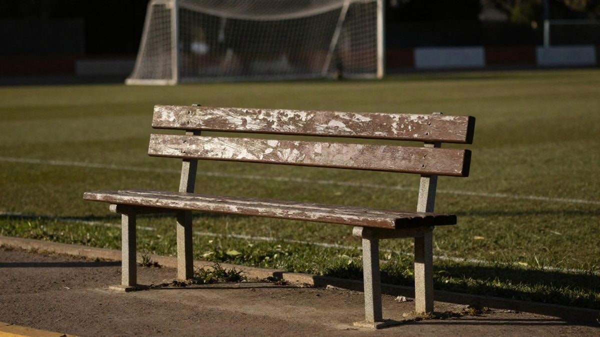 Imagen ilustrativa: banquillo de madera desgastado en el borde de un campo de fútbol, con la red de la portería al fondo