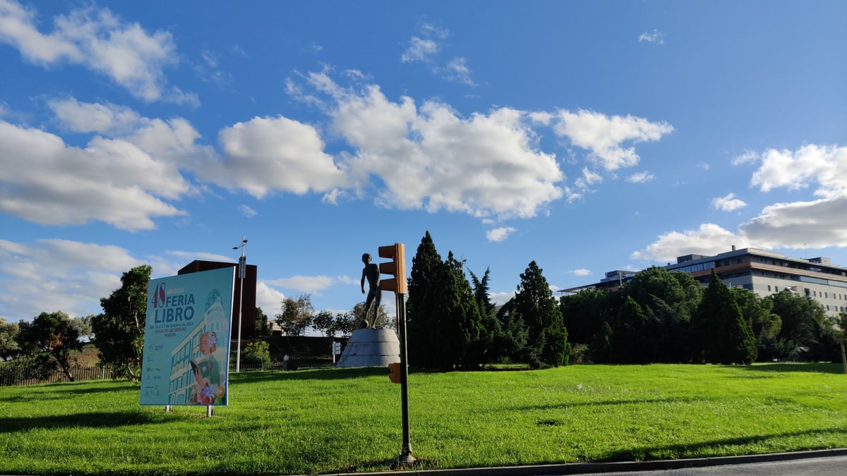 Parque con césped verde, escultura y cartel de la Feria del Libro bajo un cielo azul con nubes.