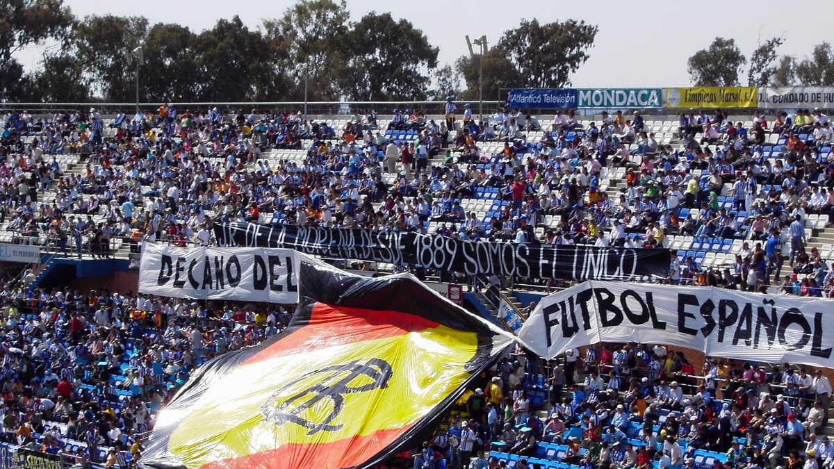 Aficionados del Recreativo de Huelva en la grada con bandera de España y pancartas azules