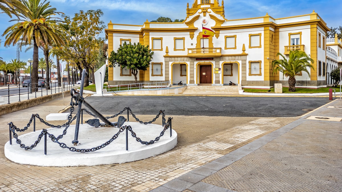 Fachada amarilla y blanca de la Comandancia de Marina con ancla y palmeras en primer plano.