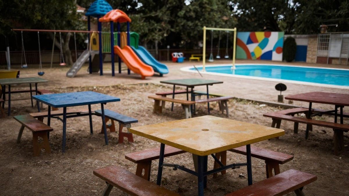 Imagen ilustrativa: un campamento de verano al aire libre con mesas de manualidades y piscina, sin personas.